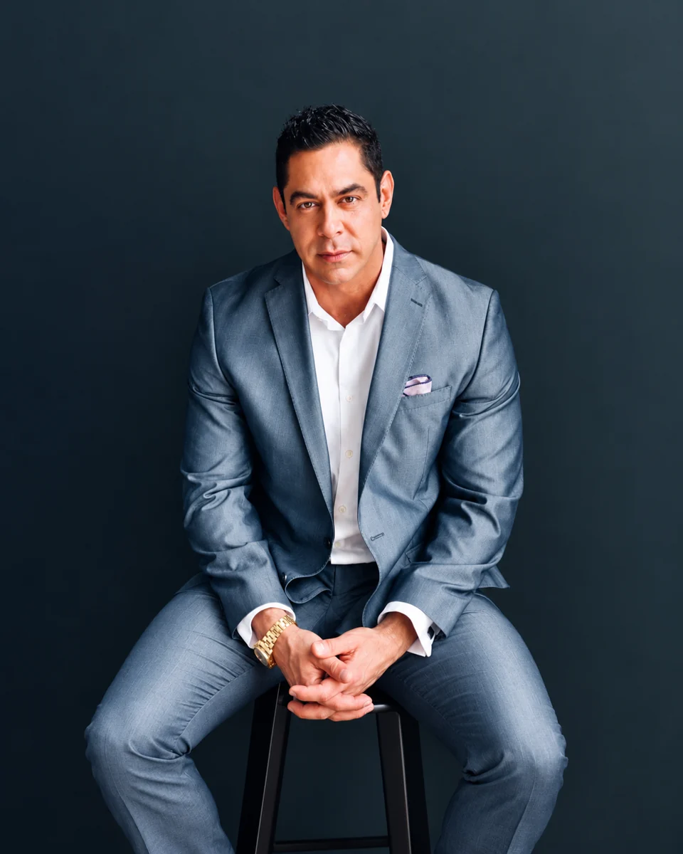 Christopher Quintana in a gray suit seated against a dark backdrop